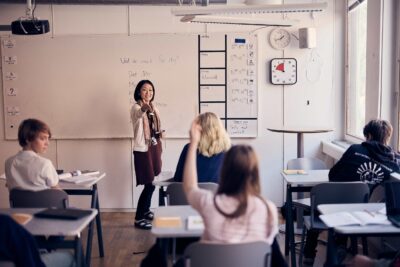 En lärare på Snitz Grundskola håller en lektion. I förgrunden sitter fyra elever, en elev räcker upp handen.